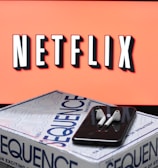 A screen displaying the Netflix logo in bold, black and white lettering against a vibrant red background. In front of the screen, there is a closed box for the board game 'Sequence', with a smartphone and wireless earbuds resting on top.